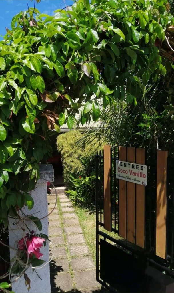 Le portillon en bois et fer forgé avec le panneau « Entrée Coco Vanille », sous la voûte de verdure