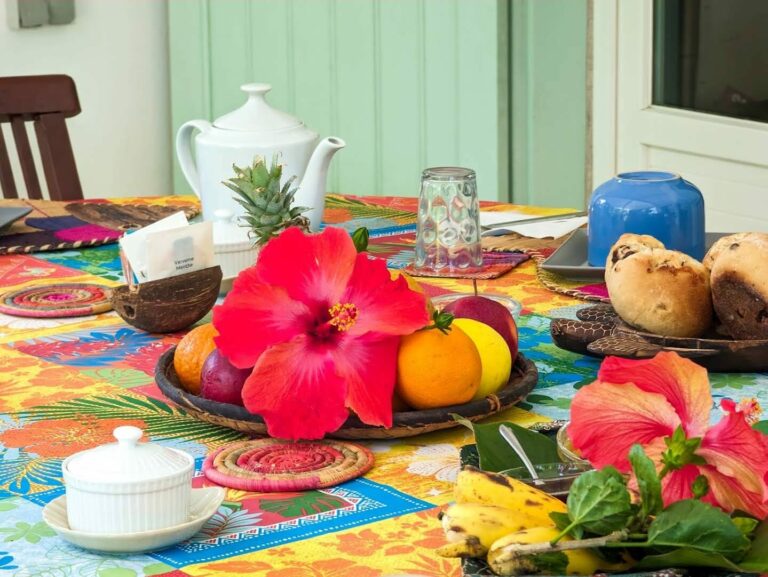 Table du petit déjeuner : théière, corbeille de fruits, hibiscus rouge et petits pains